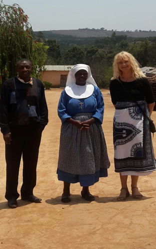 Three people standing outdoors on a dirt area, with trees and buildings in the background. The person in the middle is a woman in a blue dress and a white head covering. The person on the left is a man in dark clothing, and the person on the right is a woman with blonde hair in a black top and patterned skirt.