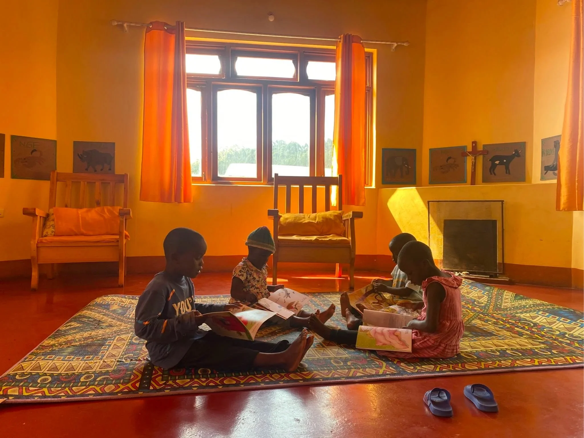 Children sitting on a colorful patterned carpet, reading books, in a room with yellow walls and a large window with orange curtains.