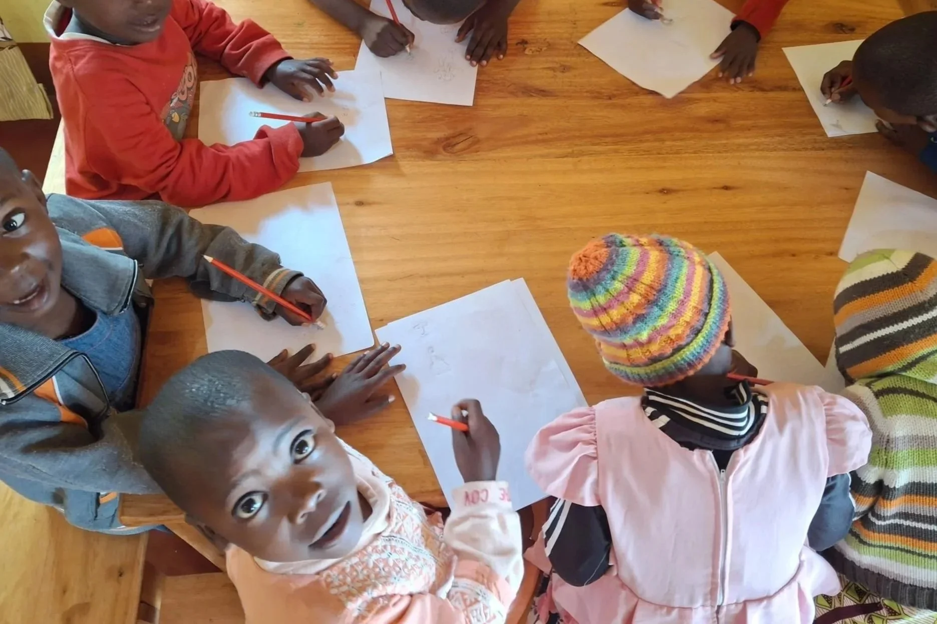 Children gathered around a wooden table, drawing on white sheets of paper with red pencils. Some children are wearing colorful hats and jackets.