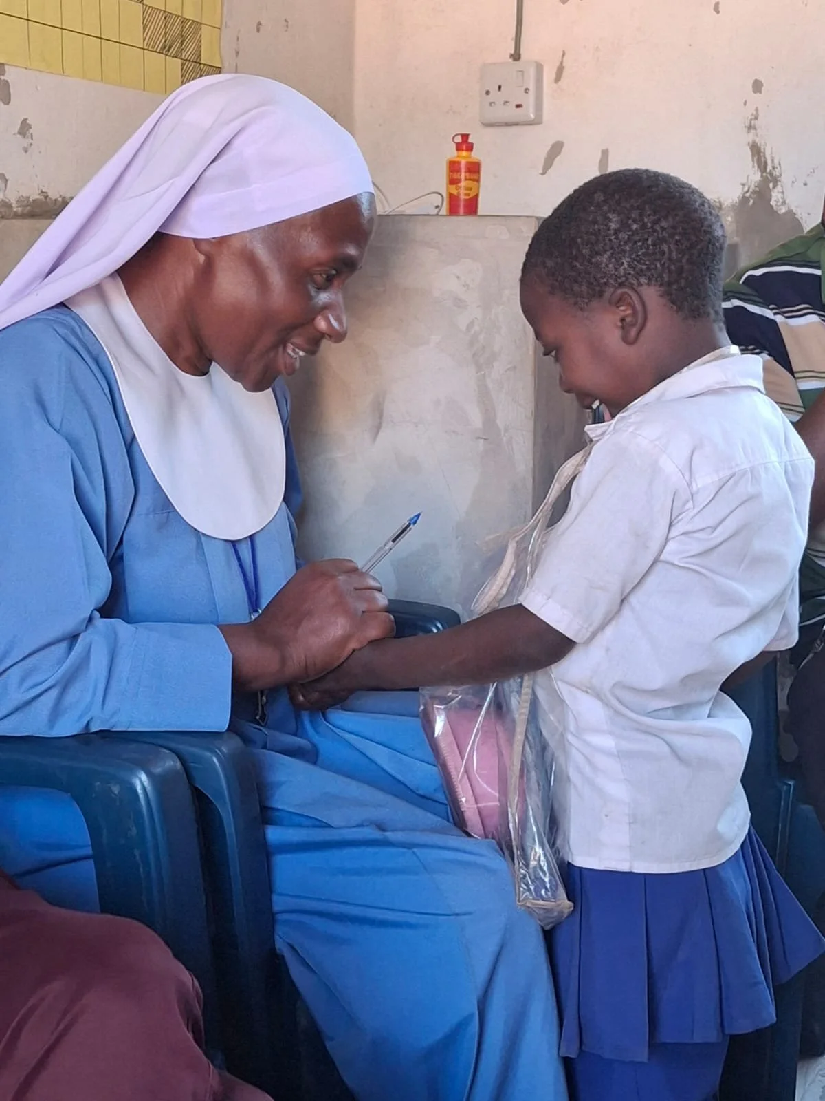 A nun sitting on a blue plastic chair, smiling and holding a pen, while a young girl in school uniform hands her a gift in a plastic bag.
