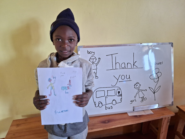 Young boy in a hoodie and beanie holding a colorful drawing, standing next to a whiteboard with thank you and drawings of a boy, girl, bus, and flower.