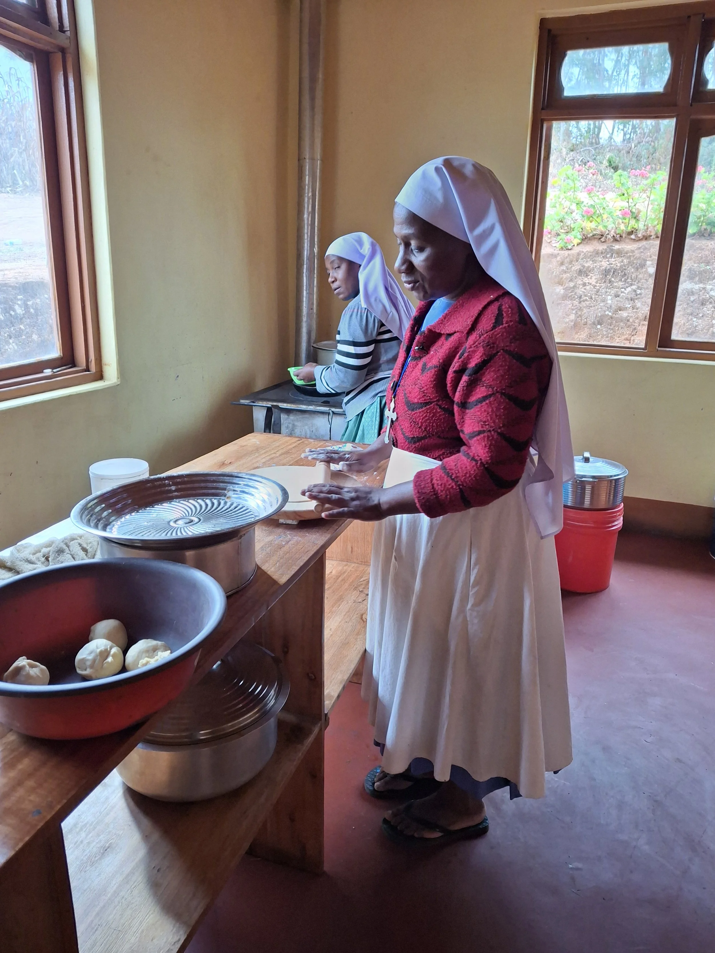 Two women wearing headscarves and aprons are preparing food in a kitchen with wooden counters. One woman is kneading dough, and there are bowls of ingredients on the counter. The room has yellow walls and open windows with views of flowers outside.