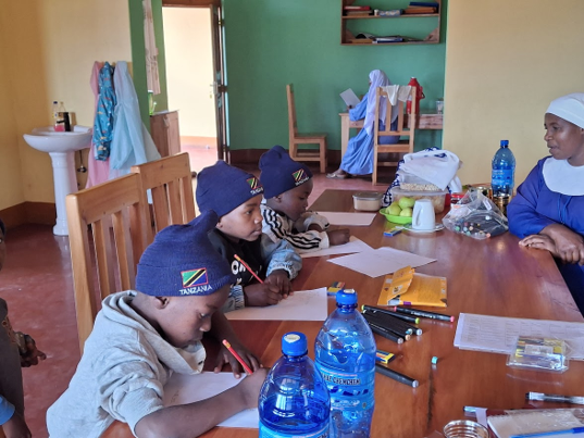 Children sitting at a wooden table, writing with pens, with a woman in a blue and white outfit in a room with yellow and green walls, and various items including bottled water, paper, and bags on the table.