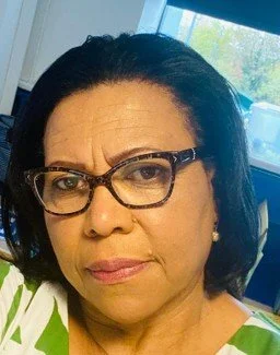 Close-up of a woman with dark hair and glasses, looking serious, in an indoor setting with a window showing outdoor trees.