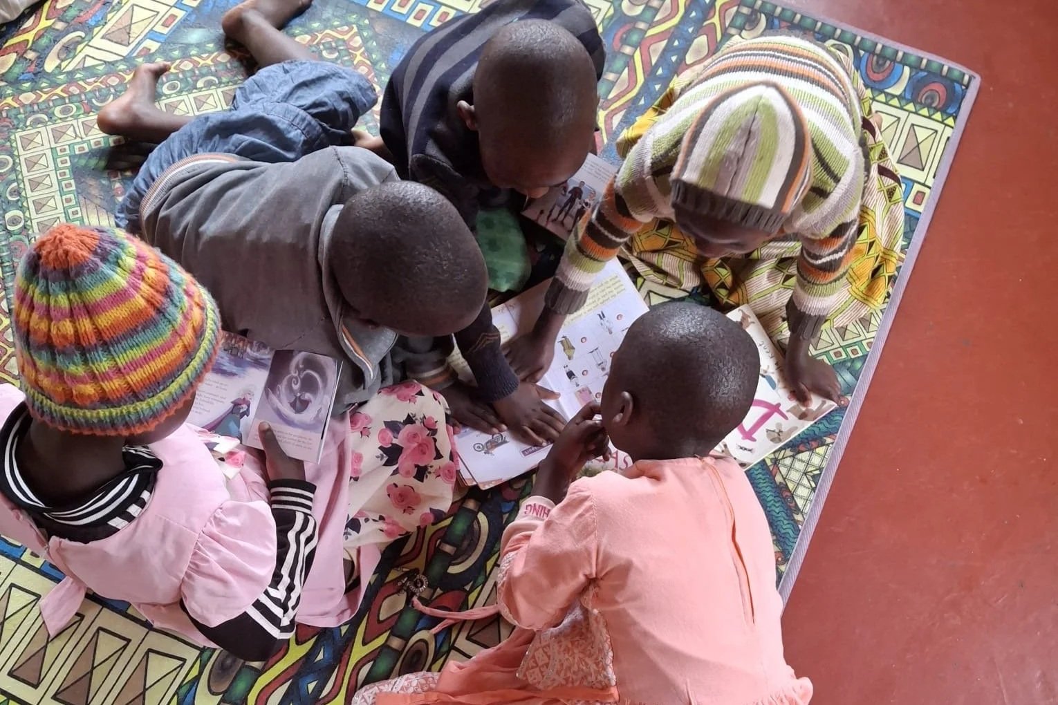 Several children are lying and sitting on a colorful patterned mat on the floor, reading books together.