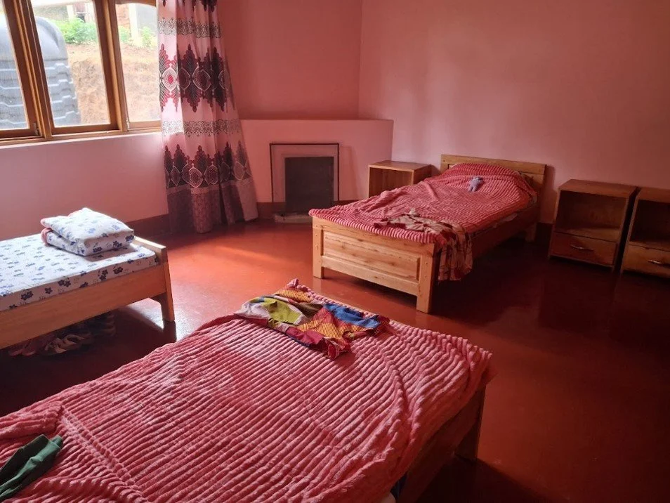 A bedroom with pink walls and wooden furniture, including two single beds with pink blankets, a nightstand, and a window with curtains. The beds have towels placed on them.