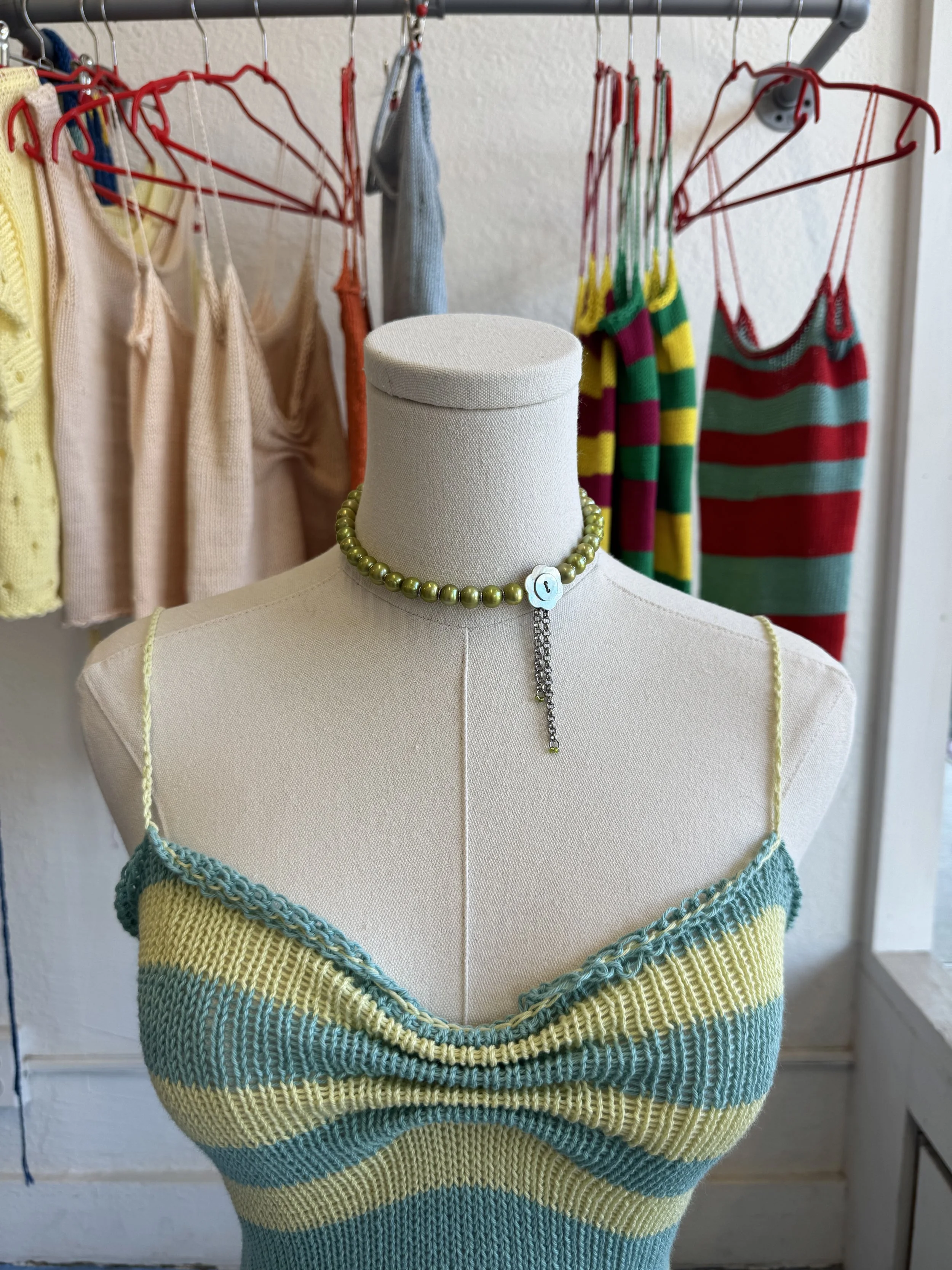 A dress form wearing a knitted tank top with yellow, green, and blue horizontal stripes, and a pearl necklace with a charm. In the background, a clothing rack with various colorful clothing items, including striped dresses and tops, hangs on red and white hangers.