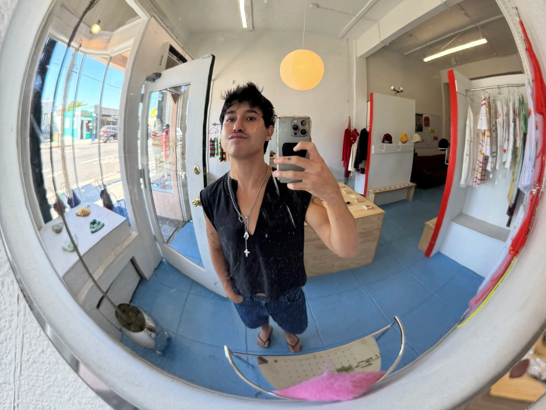 A young man taking a selfie in a round mirror inside a clothing store. The store has white walls, display racks with hats and clothing items, and outdoor scenery visible through the windows.