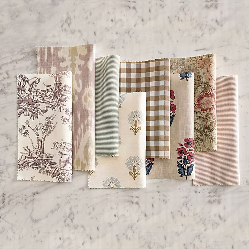 A collection of seven folded fabric swatches with various patterns and colors, arranged on a marble surface. The fabrics include floral, striped, gingham, and abstract designs in neutral tones.