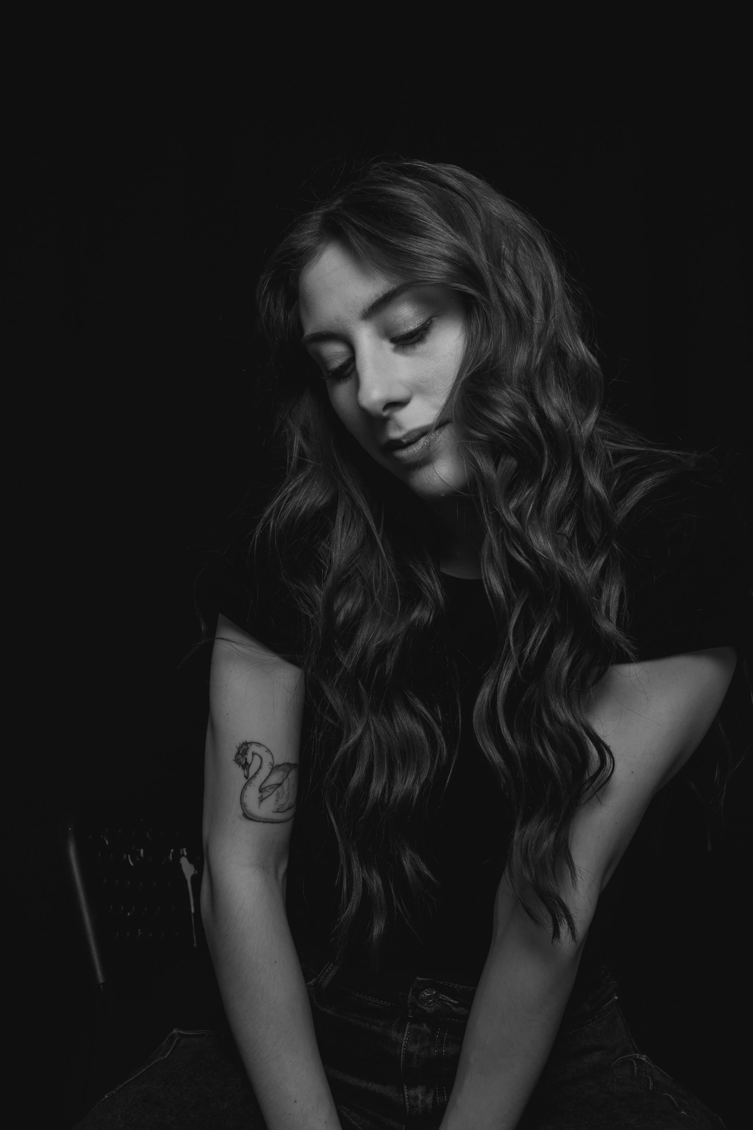 Black and white portrait of a young woman with long, curly hair and tattoo of a swan on her arm, looking down