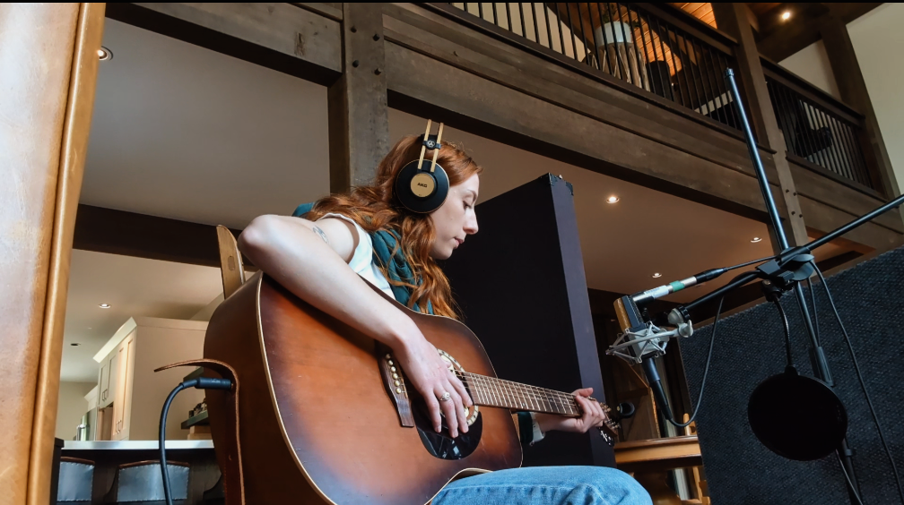 A woman with long red hair wearing headphones is playing an acoustic guitar in a cozy indoor space surrounded by professional recording equipment.