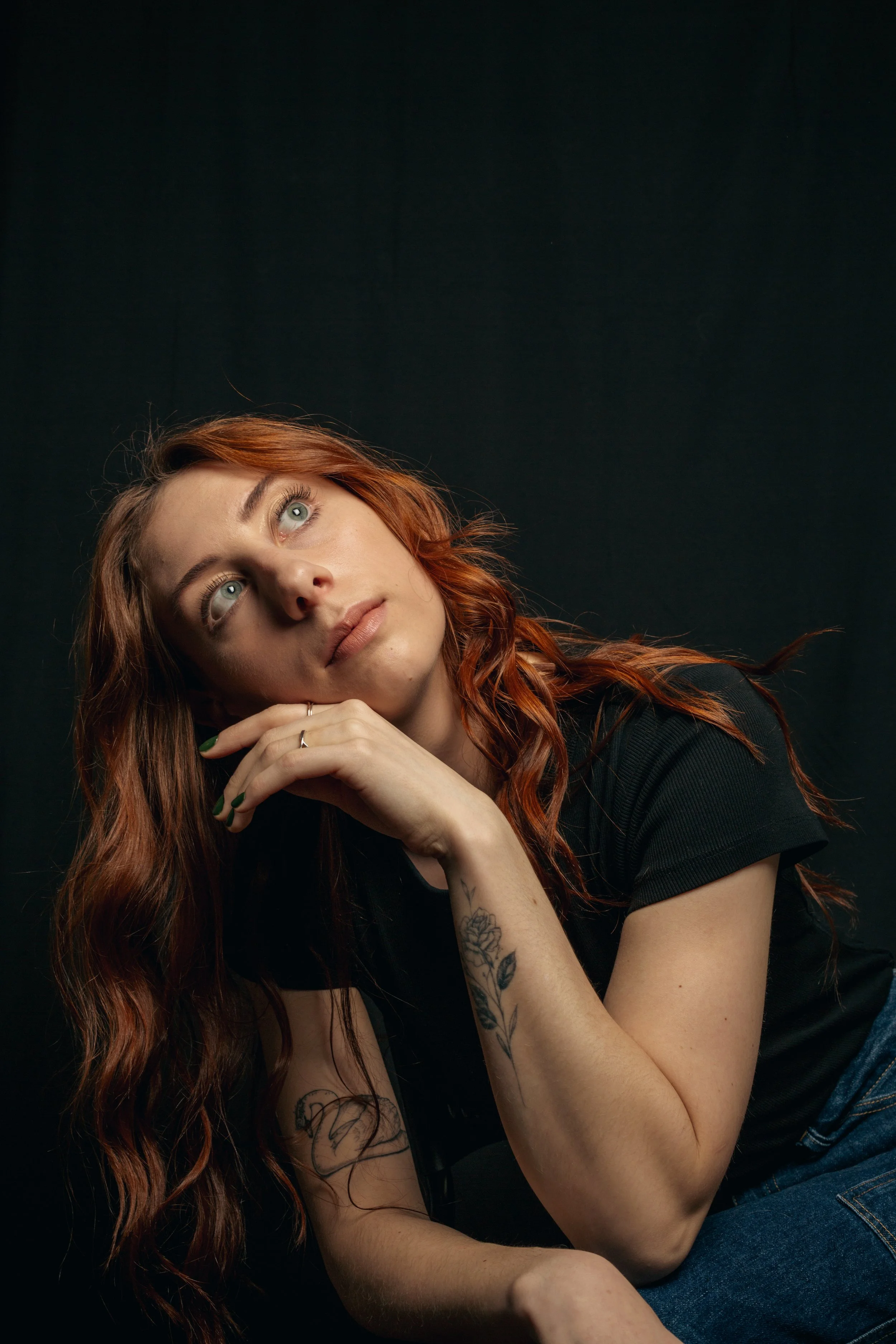 A young woman with long curly red hair and tattoos on her arm, resting her head on her hand, looking thoughtfully upward against a dark background.