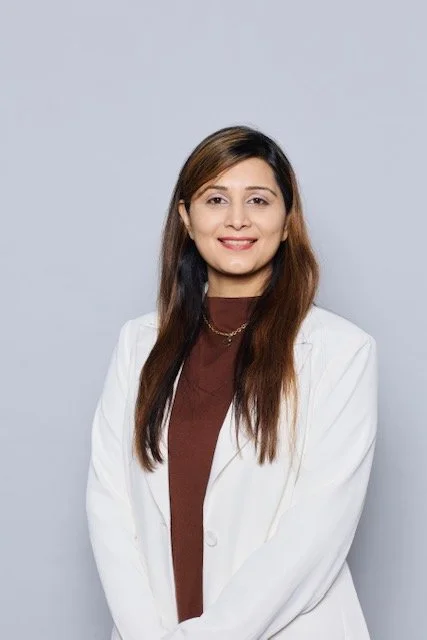A woman with long brown hair, wearing a white coat over a brown top, smiling, standing against a light gray background.