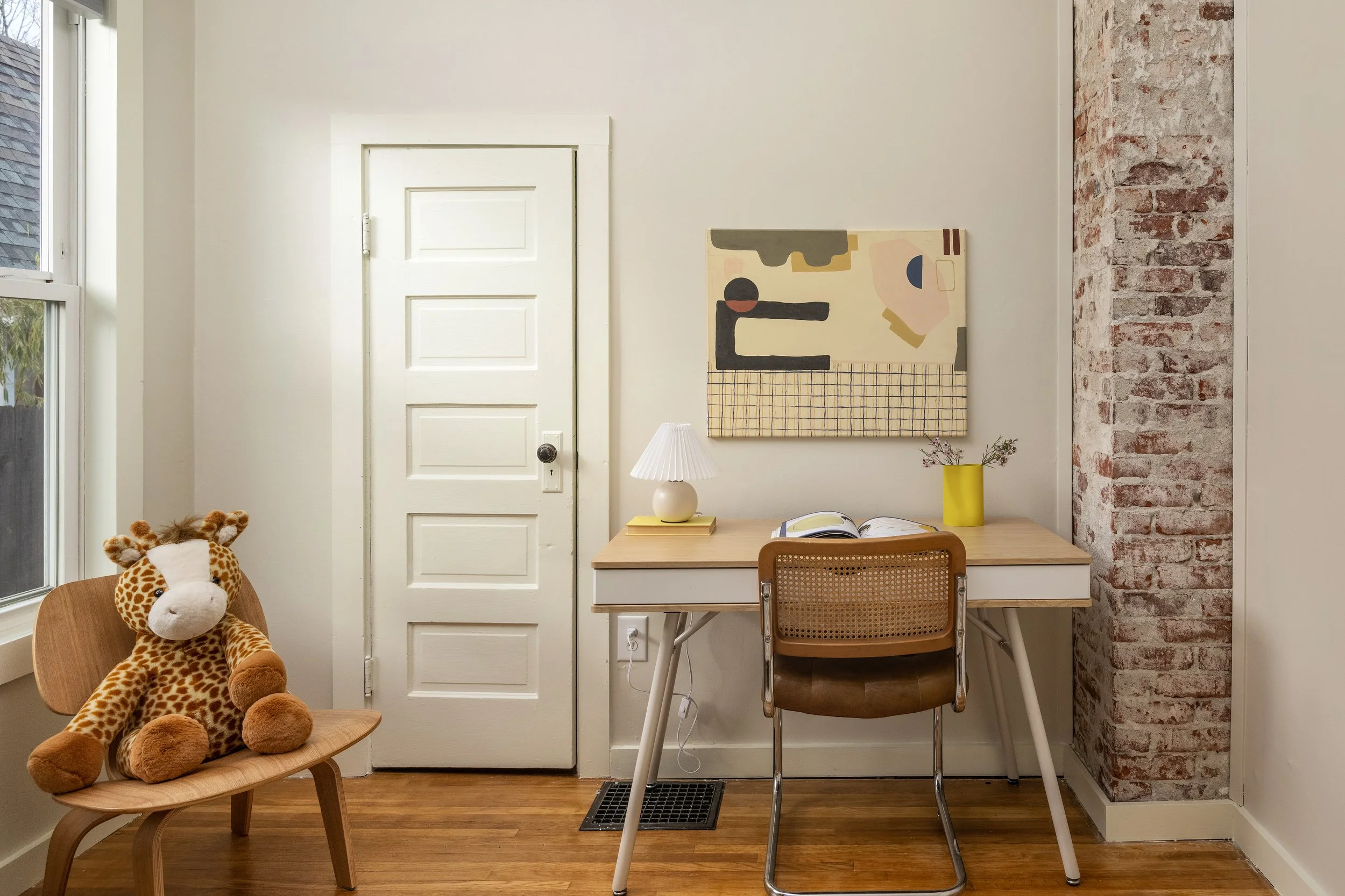 A cozy corner with a wooden rocking chair holding a stuffed giraffe toy, a small desk with an open book, a yellow vase with flowers, and a white lamp. The wall has a modern abstract art piece, a brick accent wall, and a white door. A window is visible on the left.