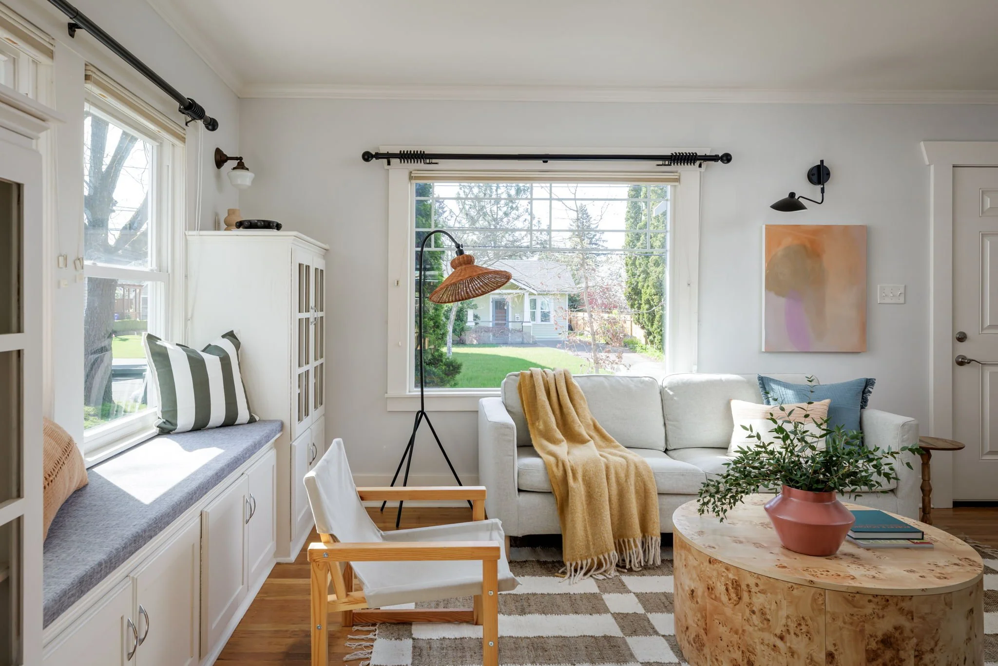 Sunlit living room with white walls, large windows, a white sofa with a yellow throw, a round marble side table with a pink vase, abstract wall art, and a window seat with black-and-white striped pillows.