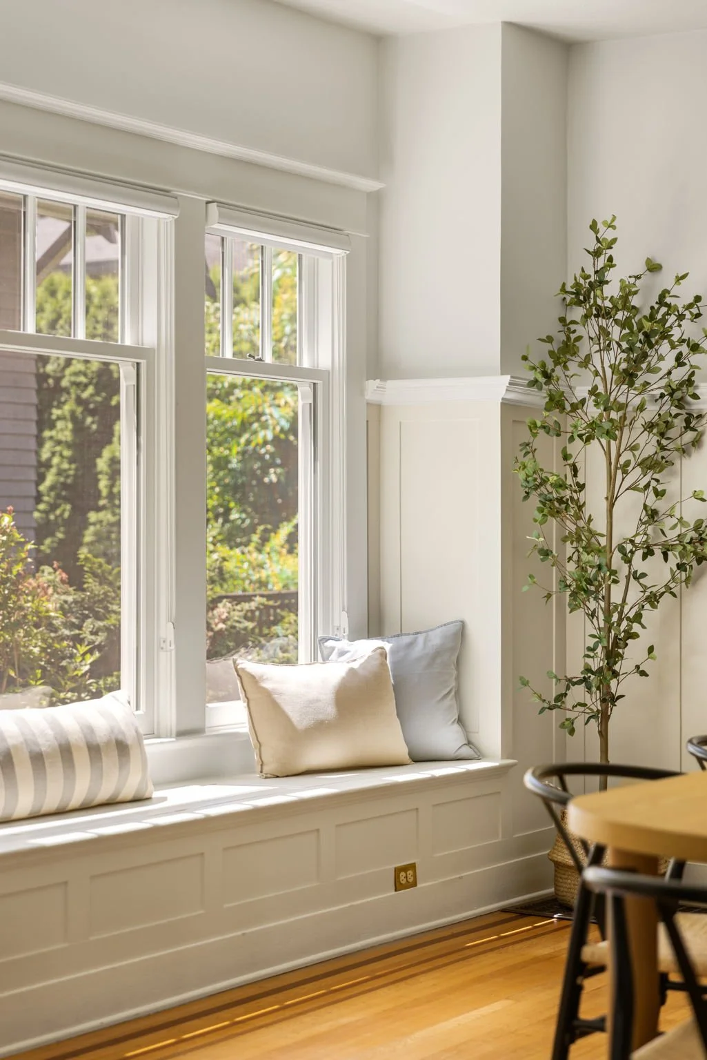 Sunlit window seat with white cushions and a striped bolster, next to large windows with views of green trees, tall potted plant in corner, wood flooring, and part of a wooden dining table with black chairs.