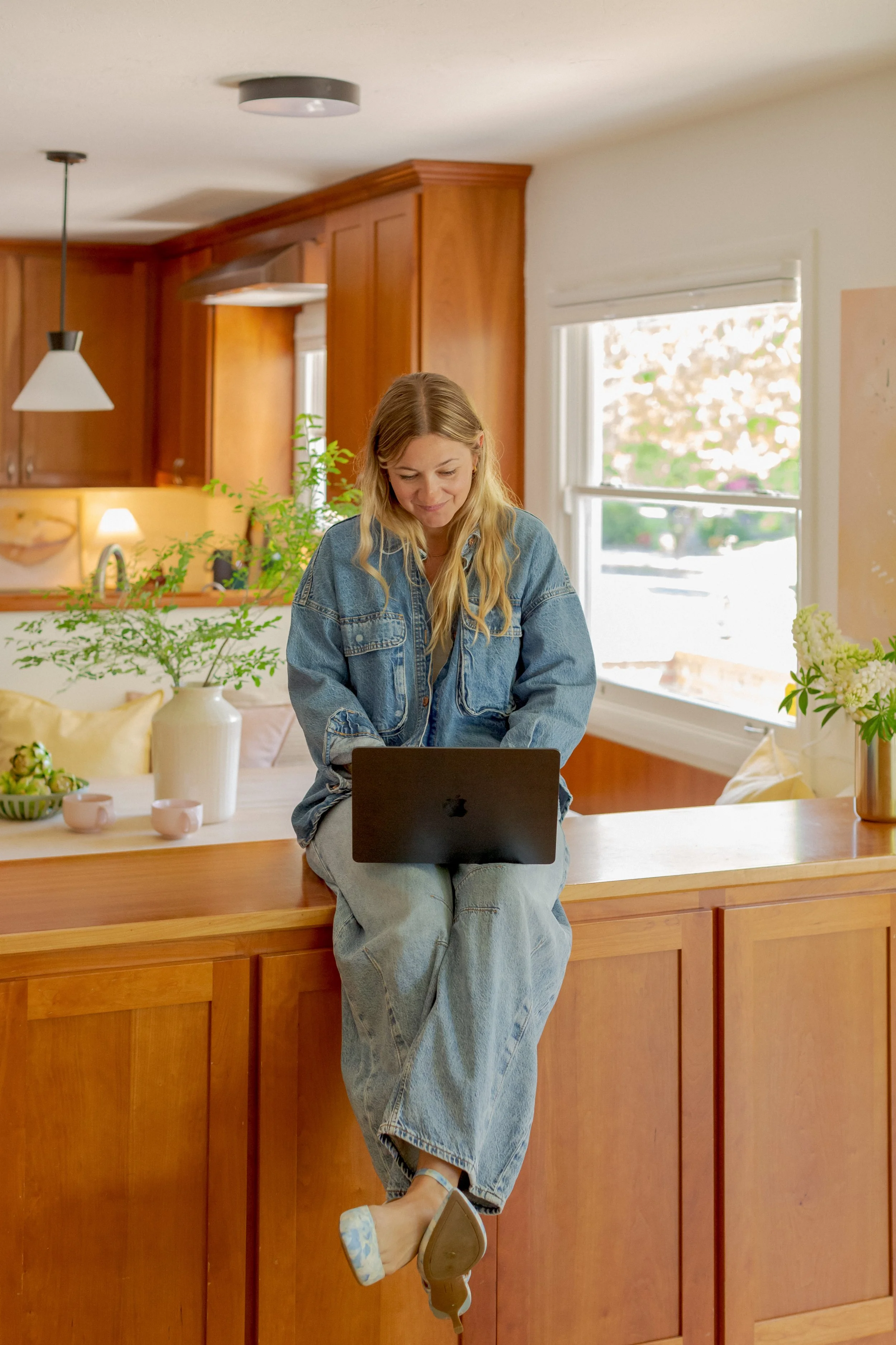 A woman with blonde hair wearing a denim jacket and light blue jeans, sitting on a kitchen counter using a laptop. The kitchen has wooden cabinets, a window with a view of trees, and decorative plants and ceramics on the counter.