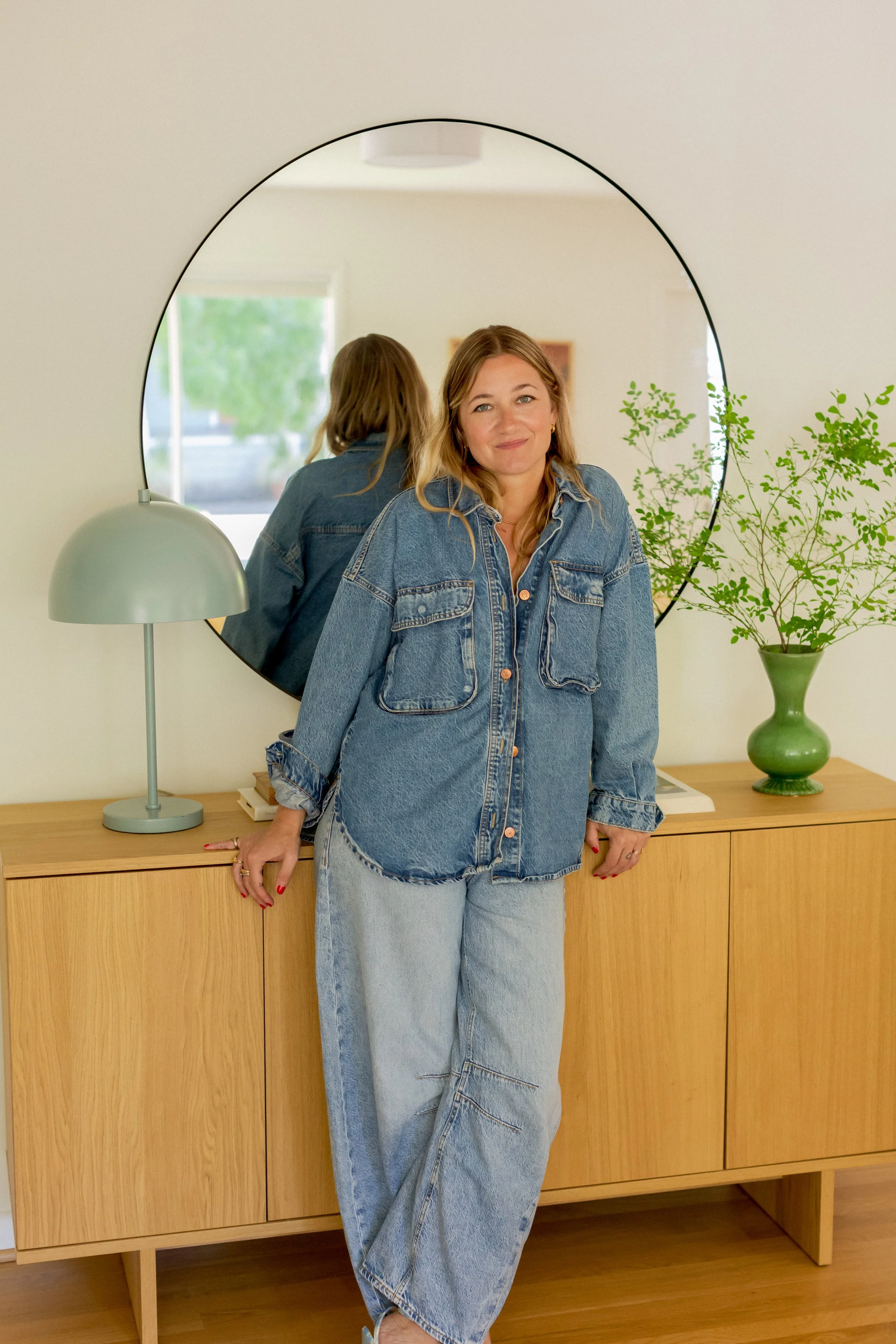 A woman in a denim jacket and jeans standing in front of a wooden sideboard with a mirror, a silver lamp, and a green vase with plants, smiling at the camera.