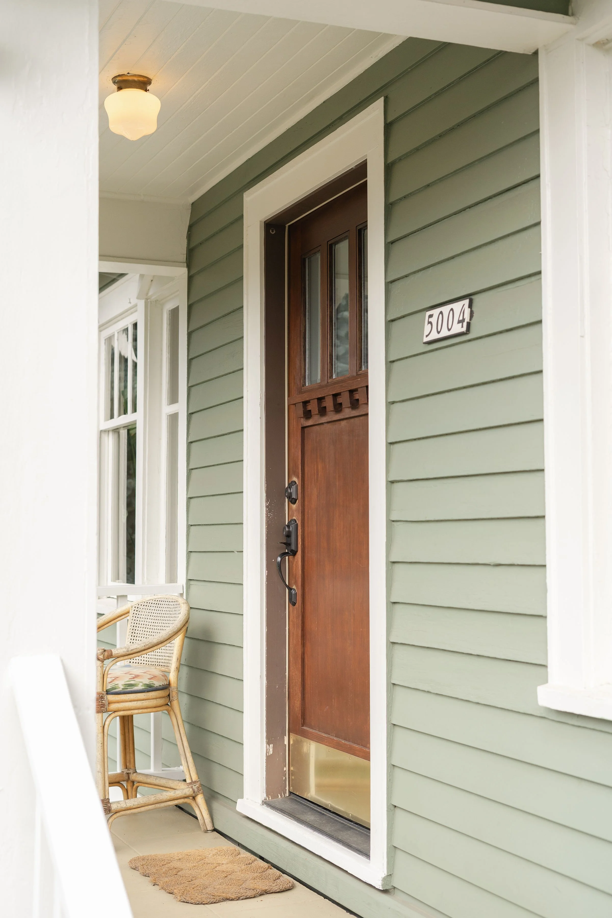 Front porch with a wooden door displaying the house number 5004, green siding, a wooden chair with a cushion, and a beige doormat, with a ceiling light fixture.