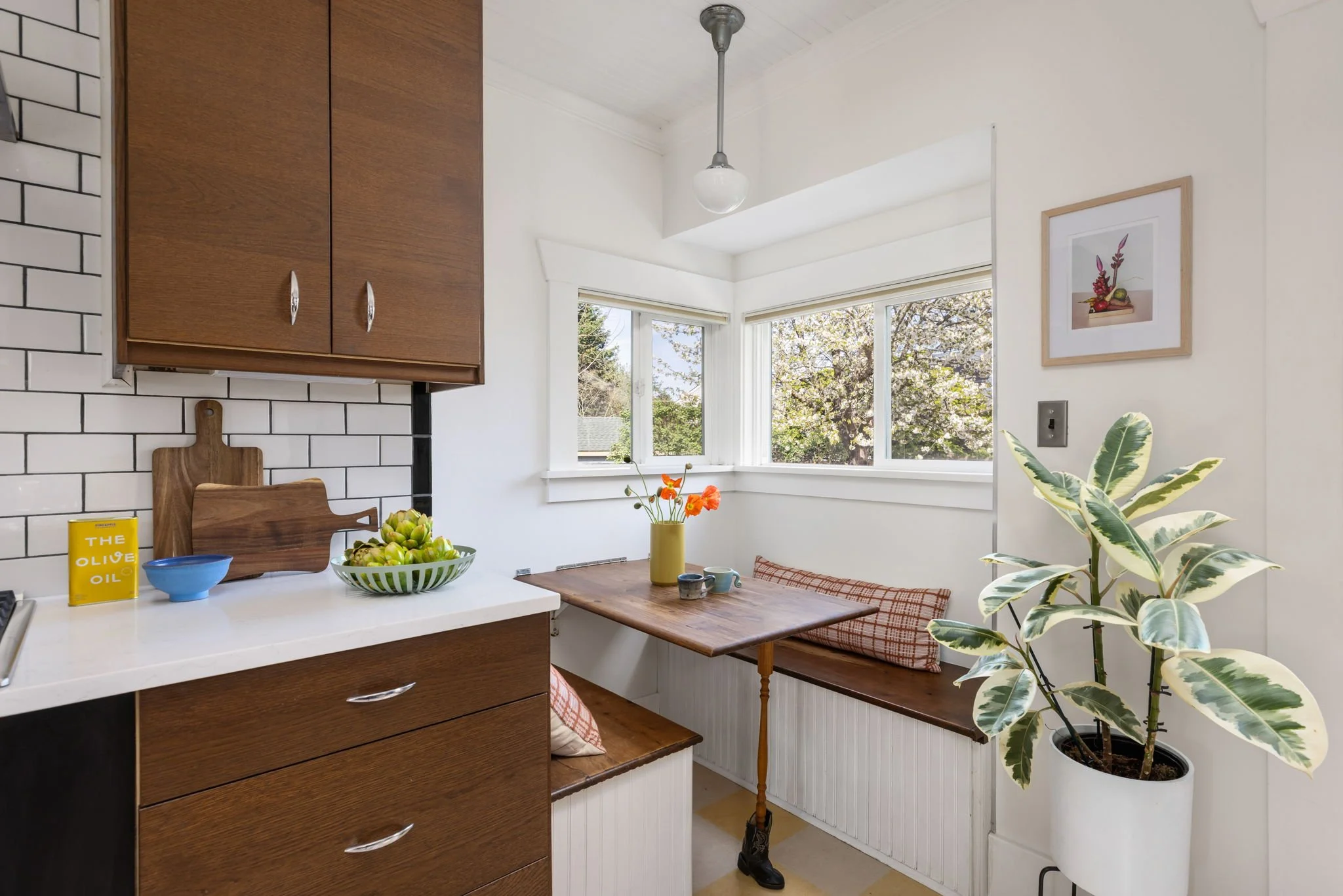 A cozy kitchen nook with a large window overlooking blooming trees. Features include white walls, a corner bench with red plaid cushions, a wooden table with a yellow flower vase, a potted plant, and framed artwork. The kitchen has a white countertop, dark wood cabinets, and a white subway tile backsplash with black grout.