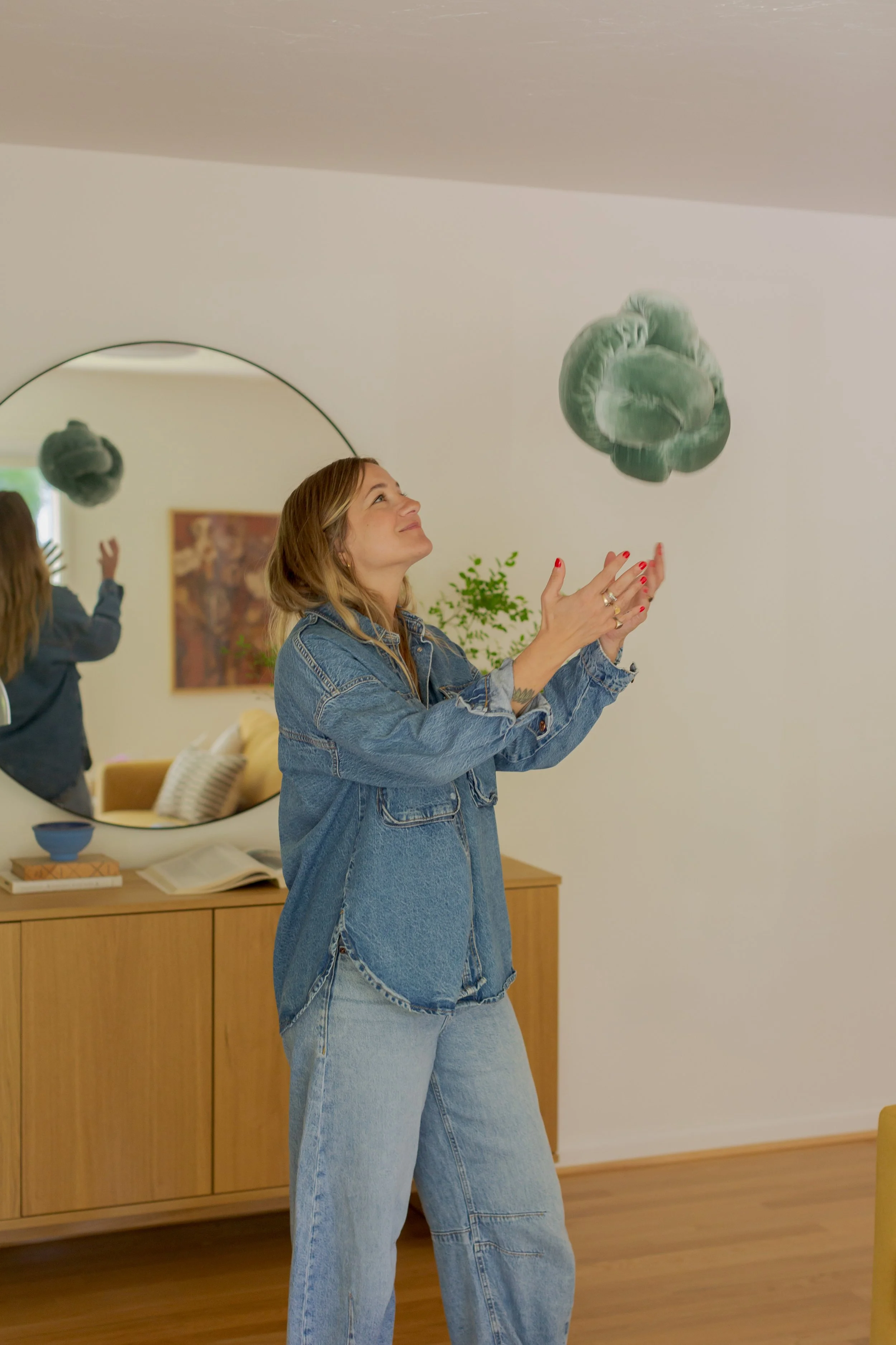A woman with blonde hair, wearing a blue denim jacket and jeans, stands in a room with wooden flooring, tossing a green knotted fabric object into the air. There is a large, round mirror on the wall reflecting her back, a wooden cabinet with books and a blue bowl on top, and a painting hanging on the wall.