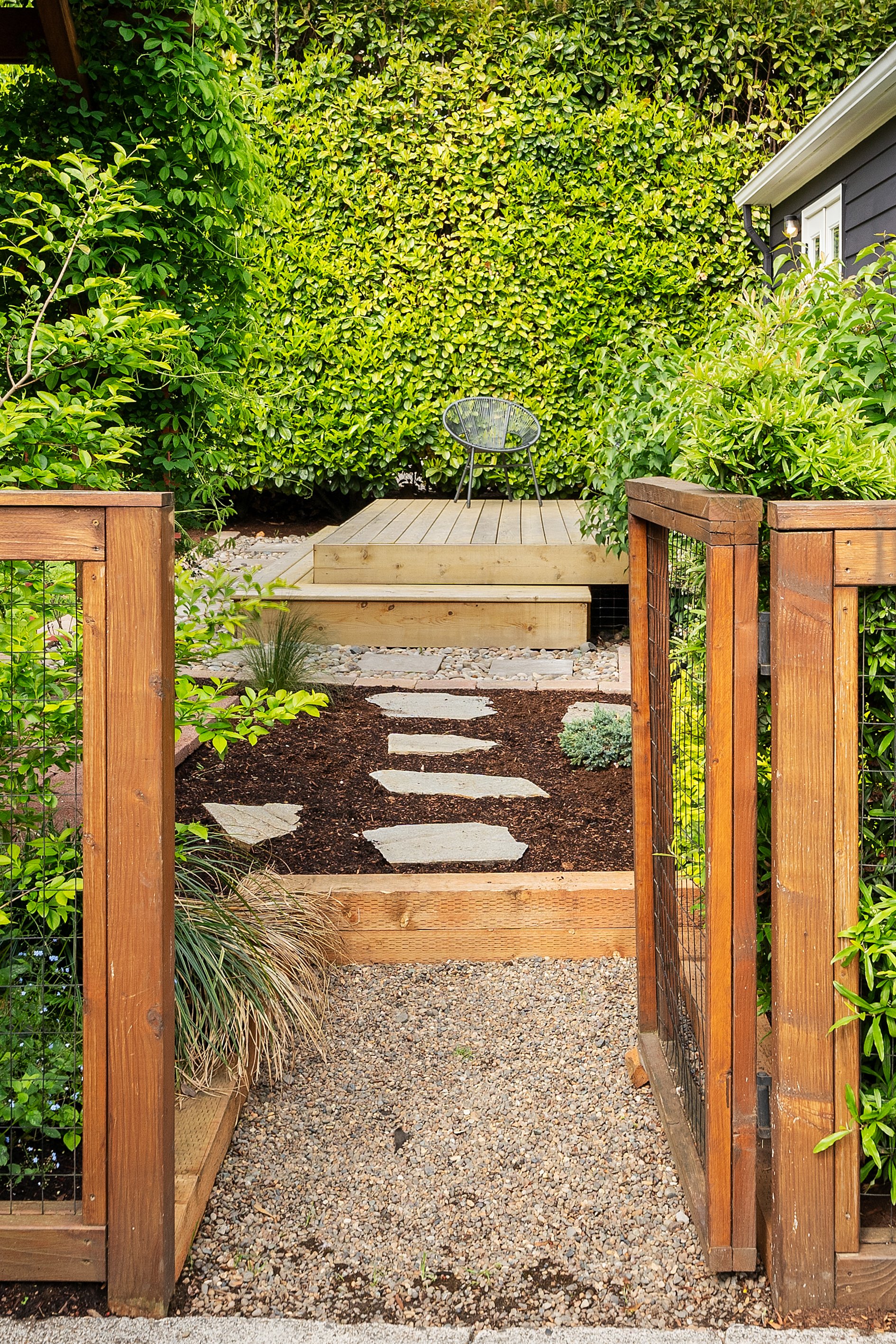 A backyard garden with a gravel pathway leading to a small wooden deck, surrounded by green bushes and plants with a house to the right.
