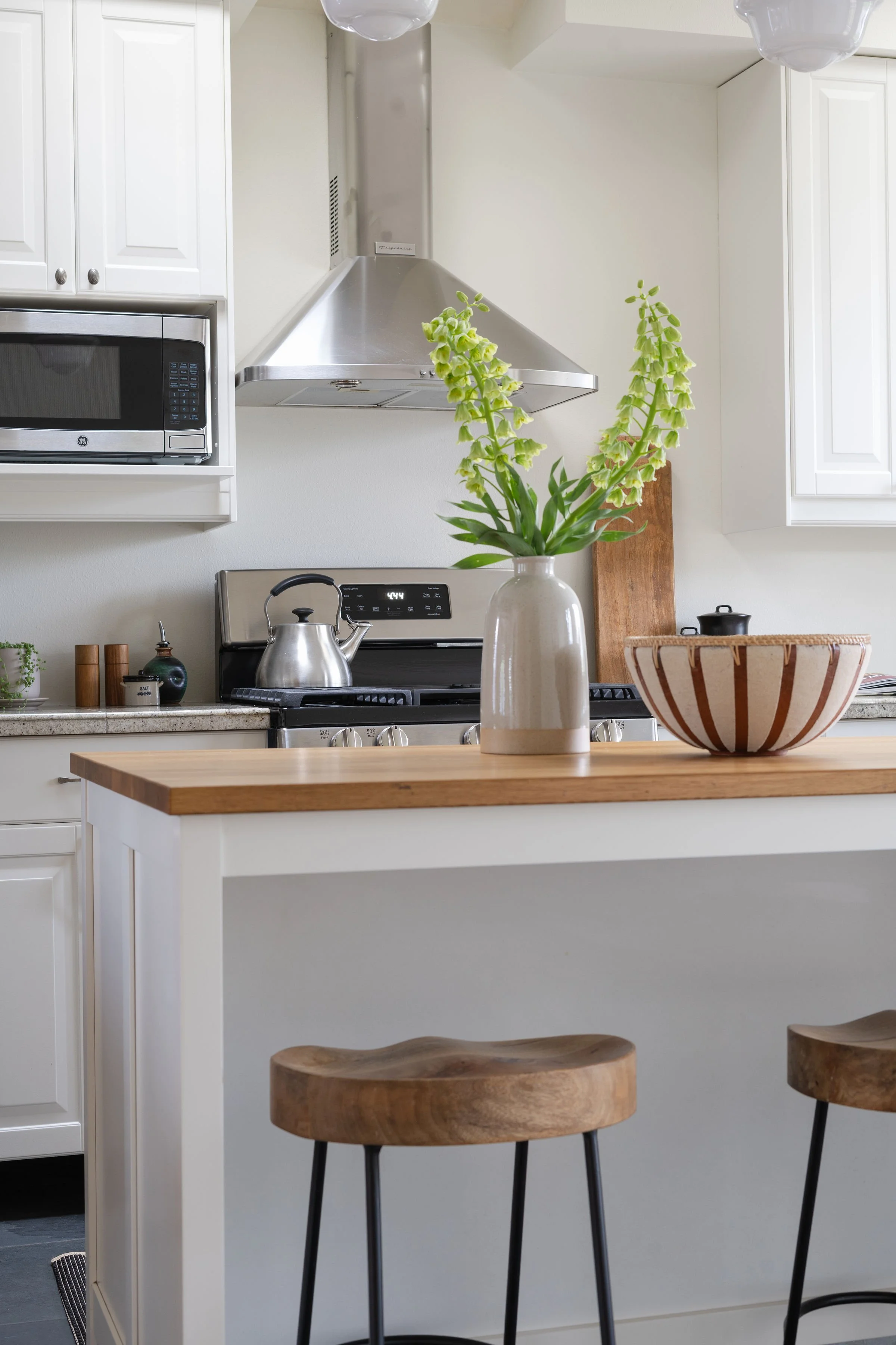 A modern kitchen with white cabinetry, a stainless steel range hood, a microwave, a stove with a kettle, and a wooden countertop with a potted plant and a striped bowl.