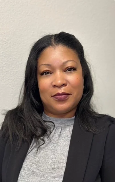 A woman with shoulder-length black hair, wearing a gray top and a black blazer, standing against a plain wall.