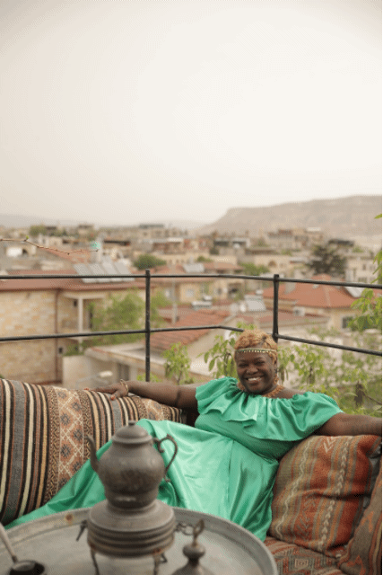 A smiling woman in a teal dress sitting on a sofa on a rooftop terrace with a city view in the background.