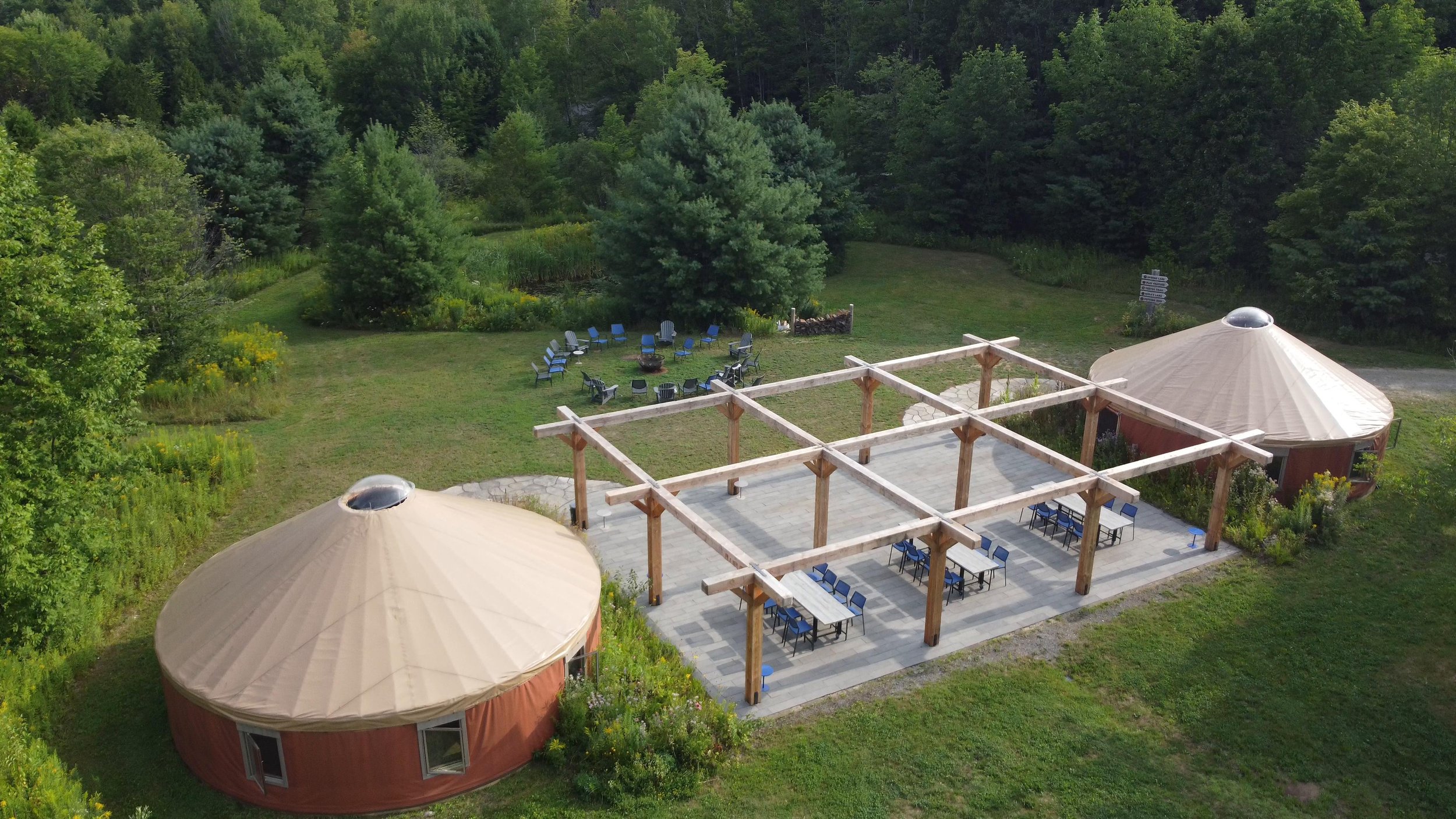Aerial view of a backyard with two round yurts, a wooden patio area with tables and chairs, a fire pit with chairs, and lush green trees surrounding the yard.