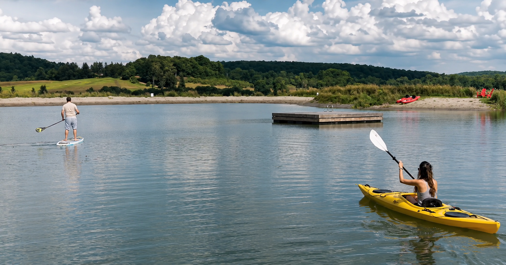 A woman in a kayak paddling on a lake, with a man stand-up paddleboarding nearby. The background features a sandy shoreline, a dock, a kayak on the shore, and lush green hills under a partly cloudy sky.
