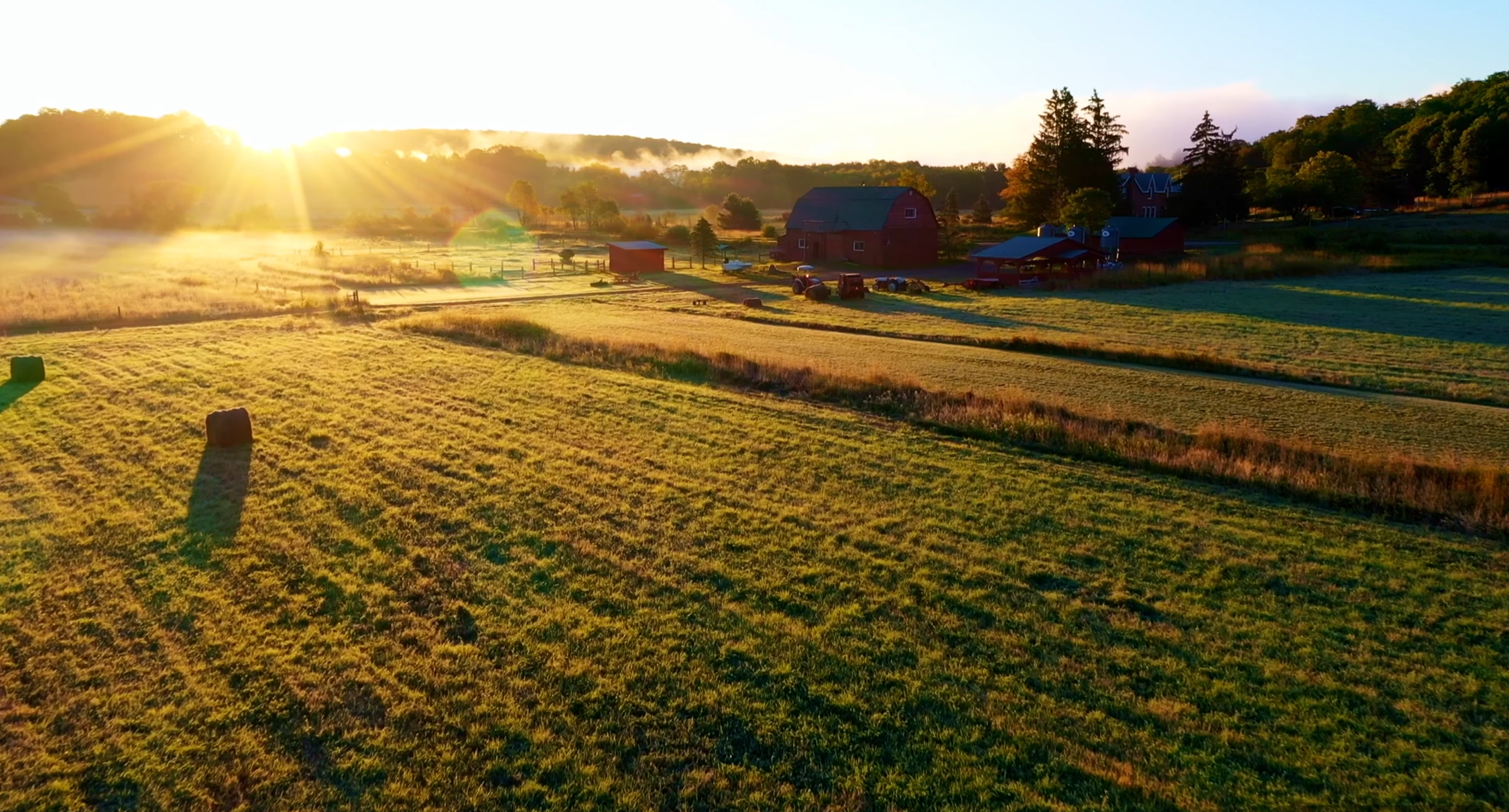 Sunrise over a farm with a red barn, trees, and hay bales in the fields.
