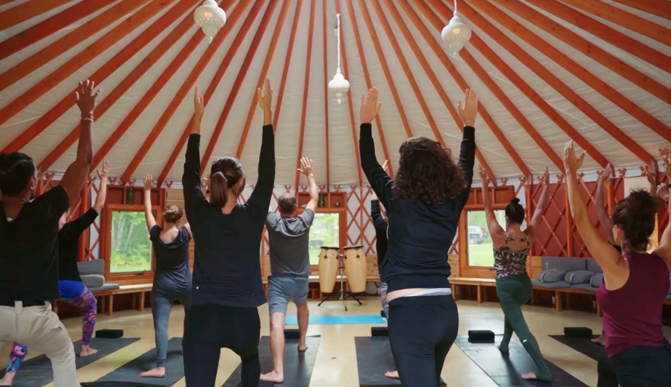 People participating in a yoga or exercise class inside a large wooden, dome-shaped structure, with some raising their hands and standing on yoga mats.