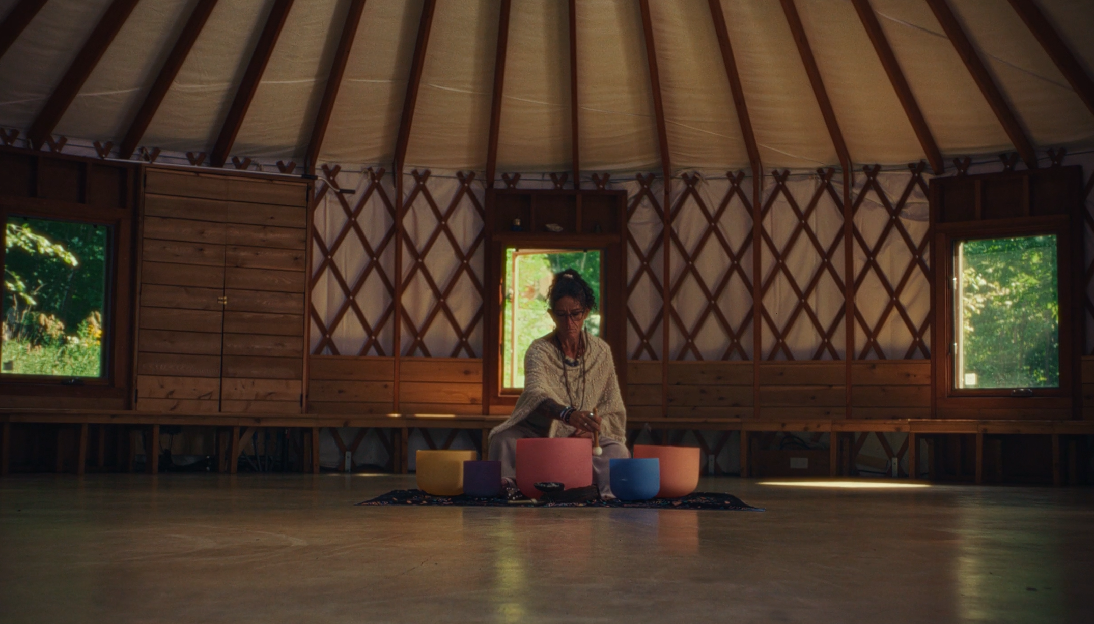 A woman sitting on a colorful rug with singing bowls inside a yurt with wooden interior and windows showing greenery outside.