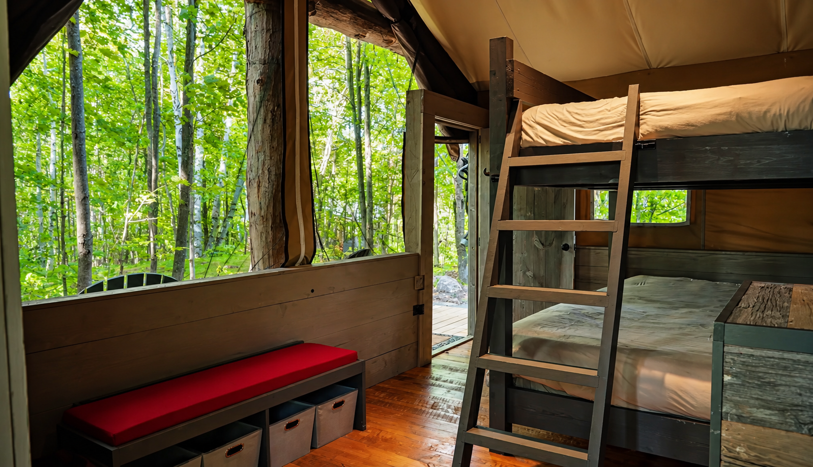 Interior of a cozy cabin with bunk beds, a window showing a lush green forest outside, a small bench with storage bins and a red cushion, and a door leading to the outdoors.