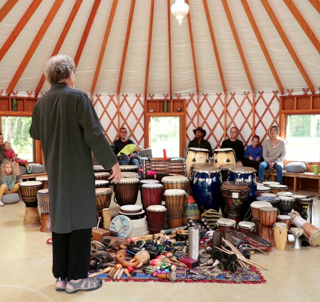 Music instructor talking to an audience in a circular music studio with rim and percussion instruments on display, some kids seated and observing.