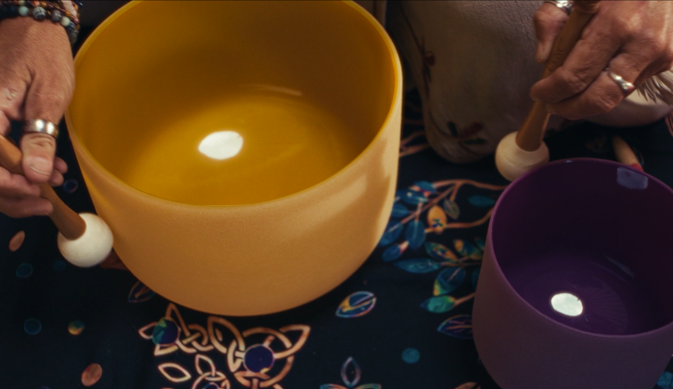 Close-up of a person's hands holding a mallet and a purple plastic cup, with a large yellow plastic cup nearby. The background features a dark tablecloth with colorful confetti and intertwined designs.