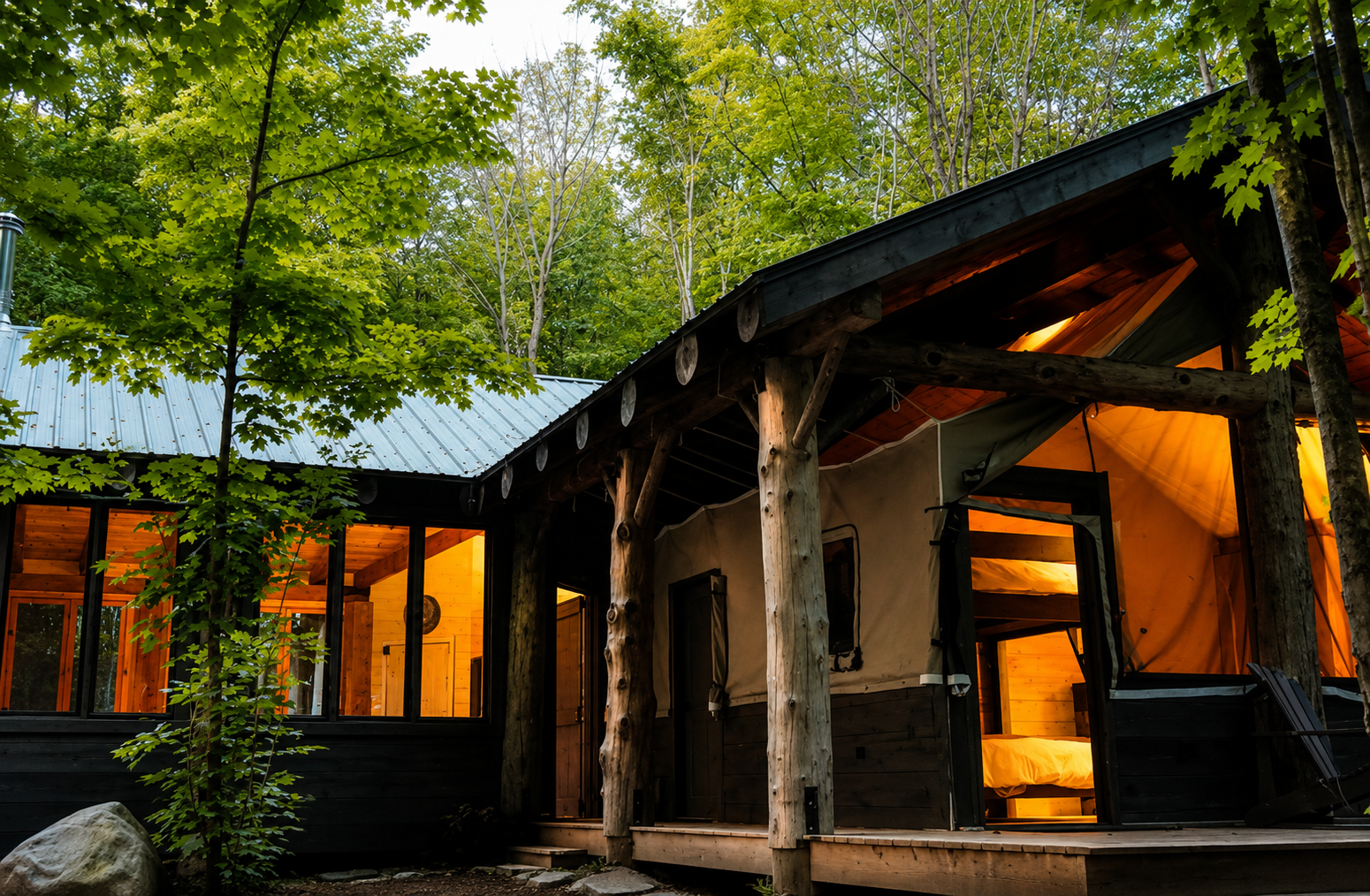 A cozy cabin with a wooden exterior and large windows surrounded by lush green trees, illuminated from inside, at dusk.