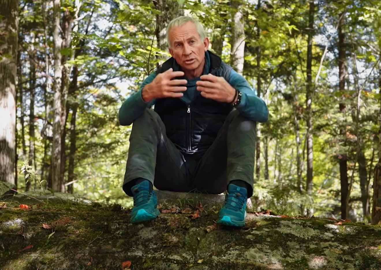 An older man with gray hair sitting on a moss-covered log in a dense forest, wearing outdoor clothing and blue hiking shoes, speaking and gesturing with his hands.