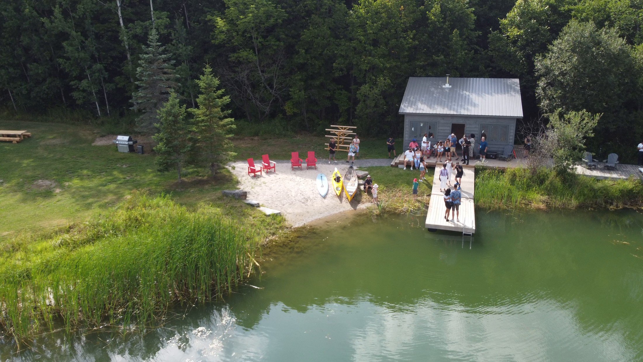 People gathering outside a small gray building by a lake, with red chairs, kayaks, and a dock, surrounded by trees.