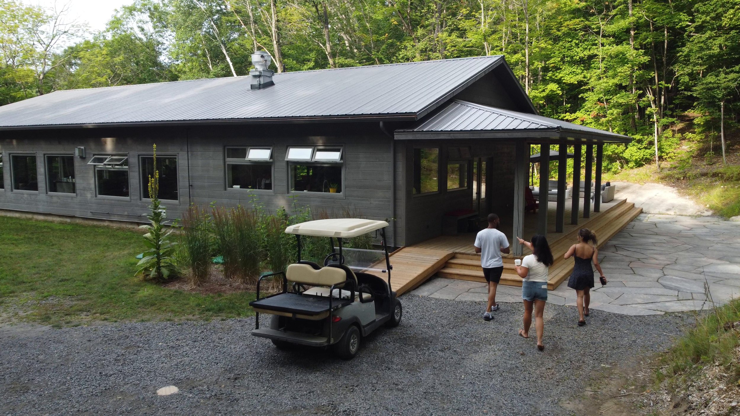 A modern gray house with a metal roof, surrounded by trees, with a small wooden deck and people walking on a pathway in front.