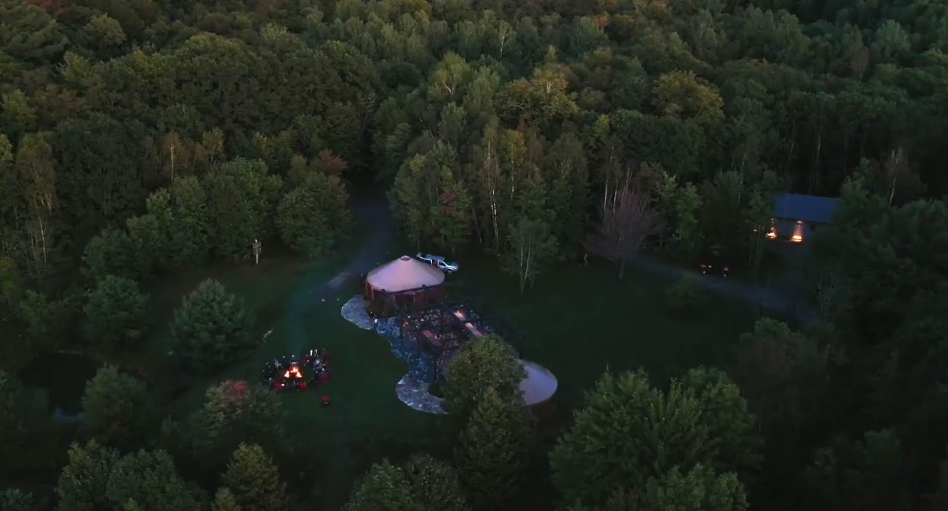An aerial view of a wooded area at dusk featuring a circular outdoor event tent with some lighting, a small fire pit with people gathered around, parked cars, and a nearby house with lights on.