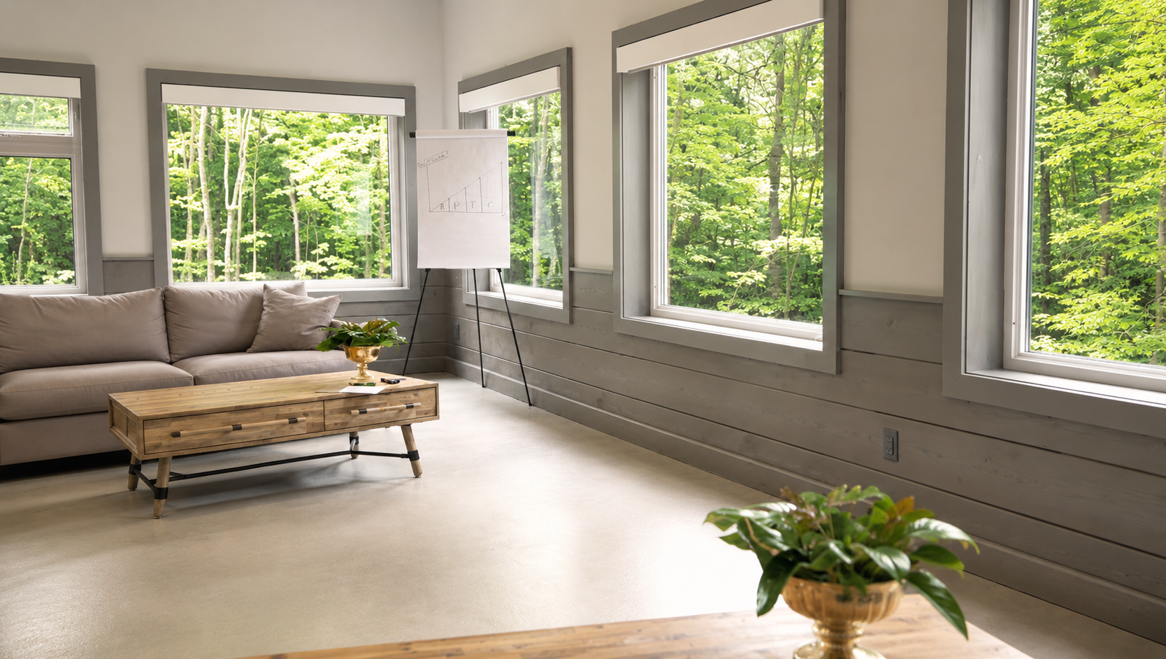 A bright living room with beige couch, wooden coffee table, large windows overlooking green trees, a whiteboard with a graph, and potted plants.