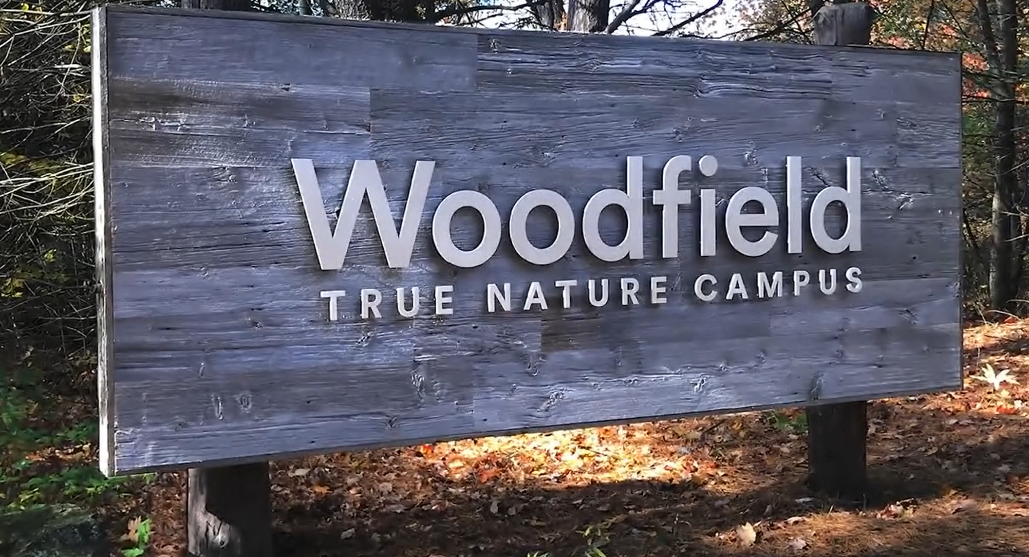 A wooden sign that reads 'Woodfield True Nature Campus' with a background of trees and fallen leaves.
