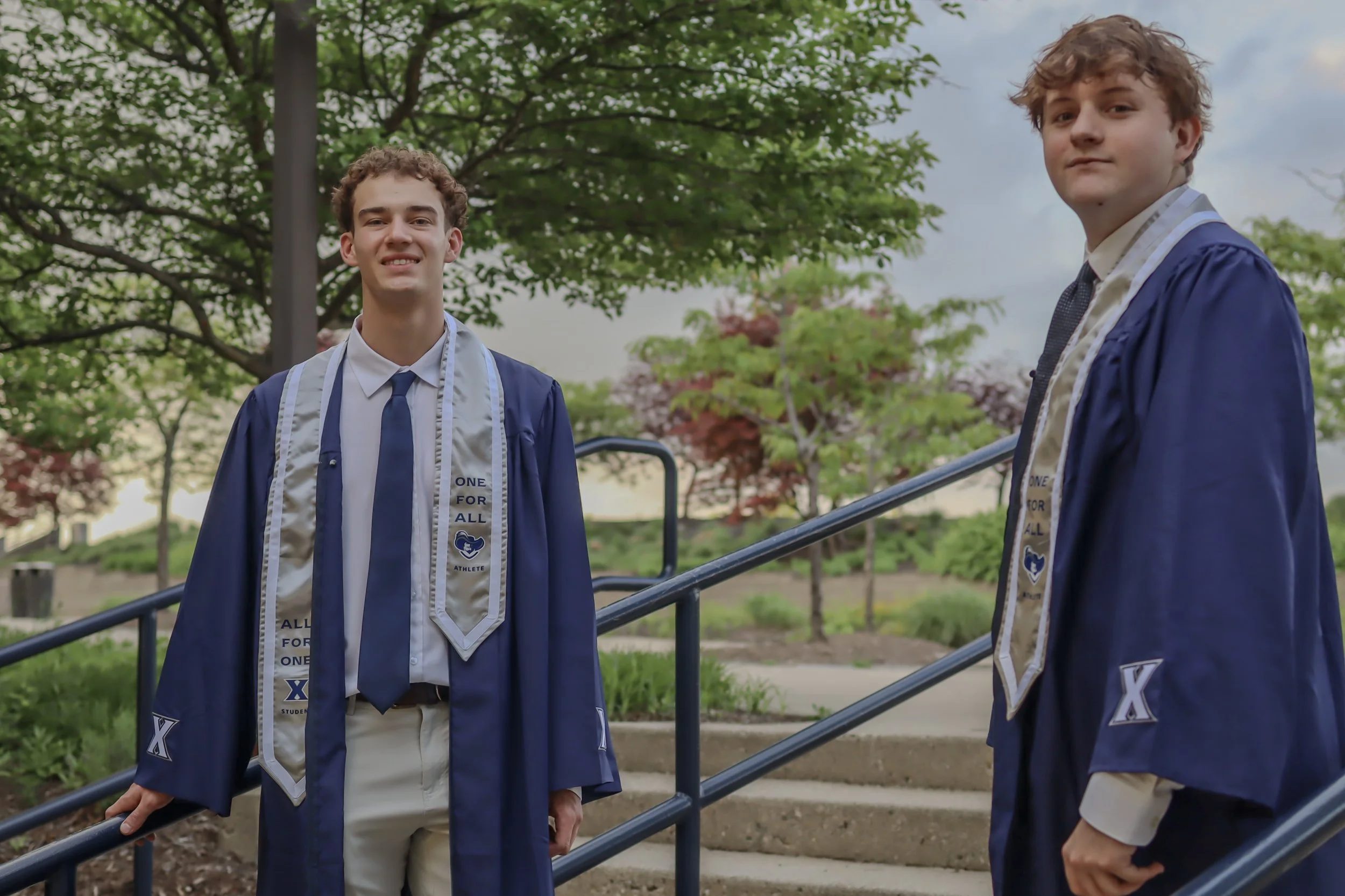 Two young men wearing blue graduation gowns and white sashes with medals are standing outdoors near a blue railing, with green trees and a cloudy sky in the background.