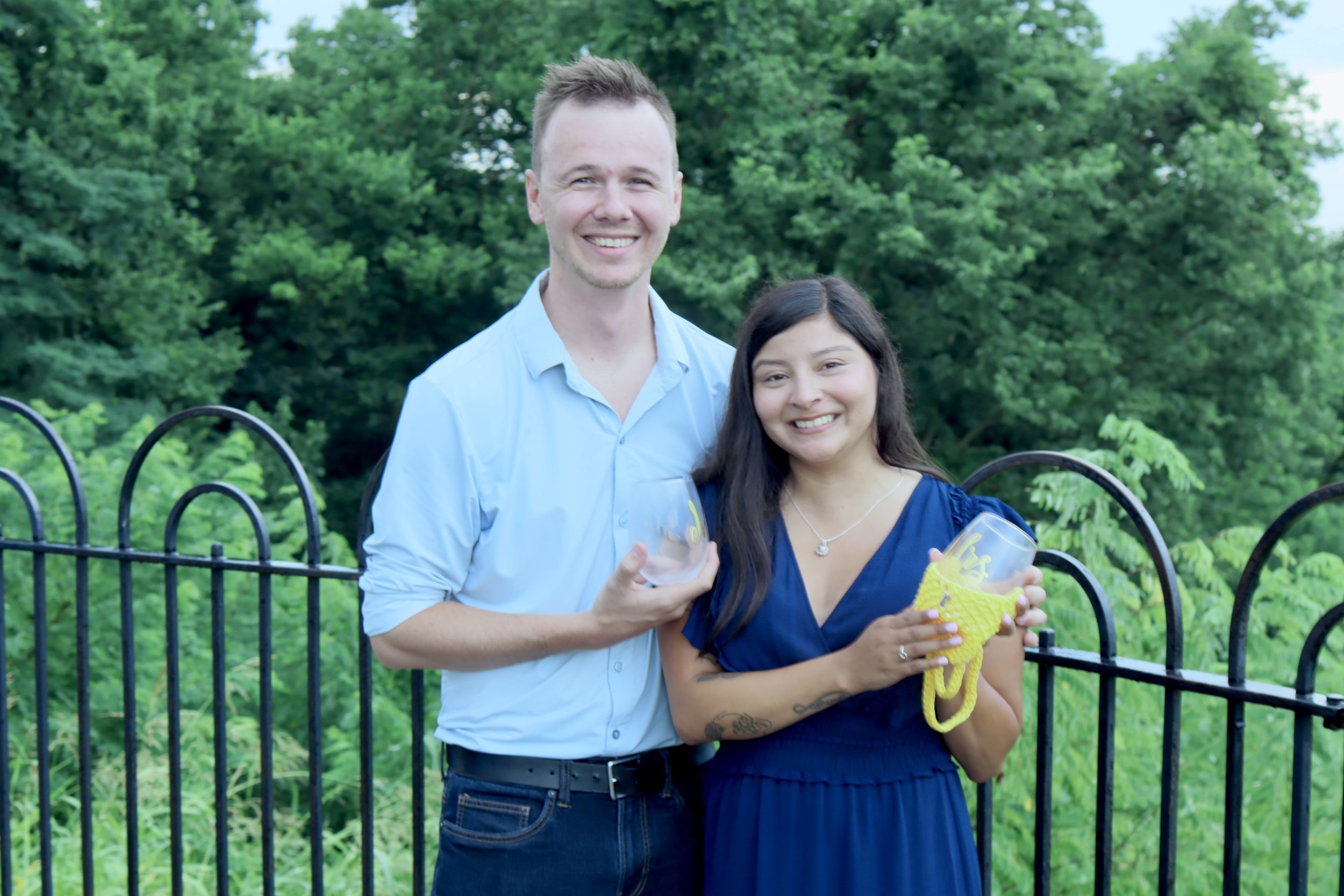 A young man and woman smiling and holding clear plastic cups with yellow and purple decorations, standing outdoors near a black metal fence with trees and greenery in the background.
