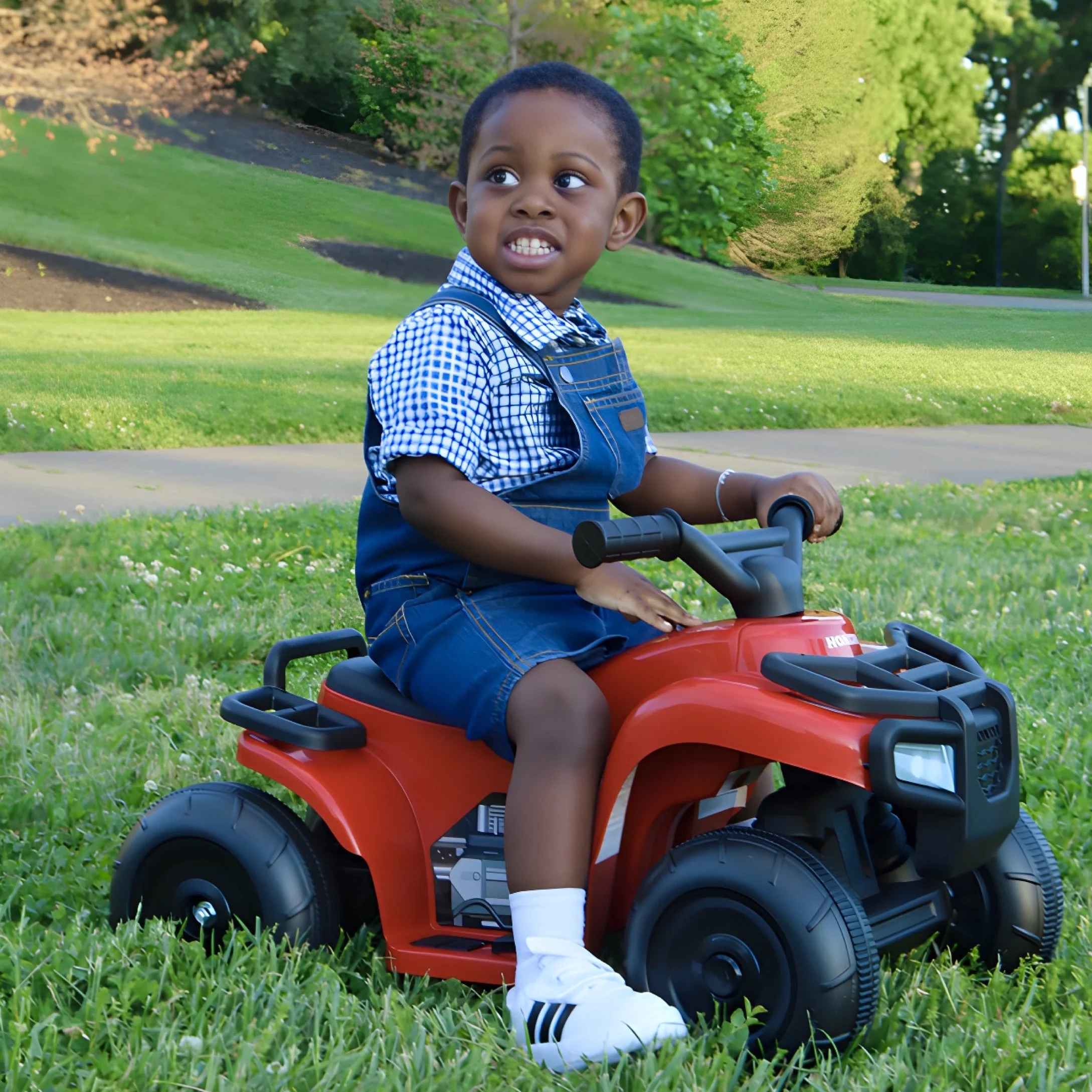 A young boy sitting on a red toy car outdoors in a park, wearing a blue and white checkered shirt, denim overalls, and white sneakers, with trees and grass in the background.