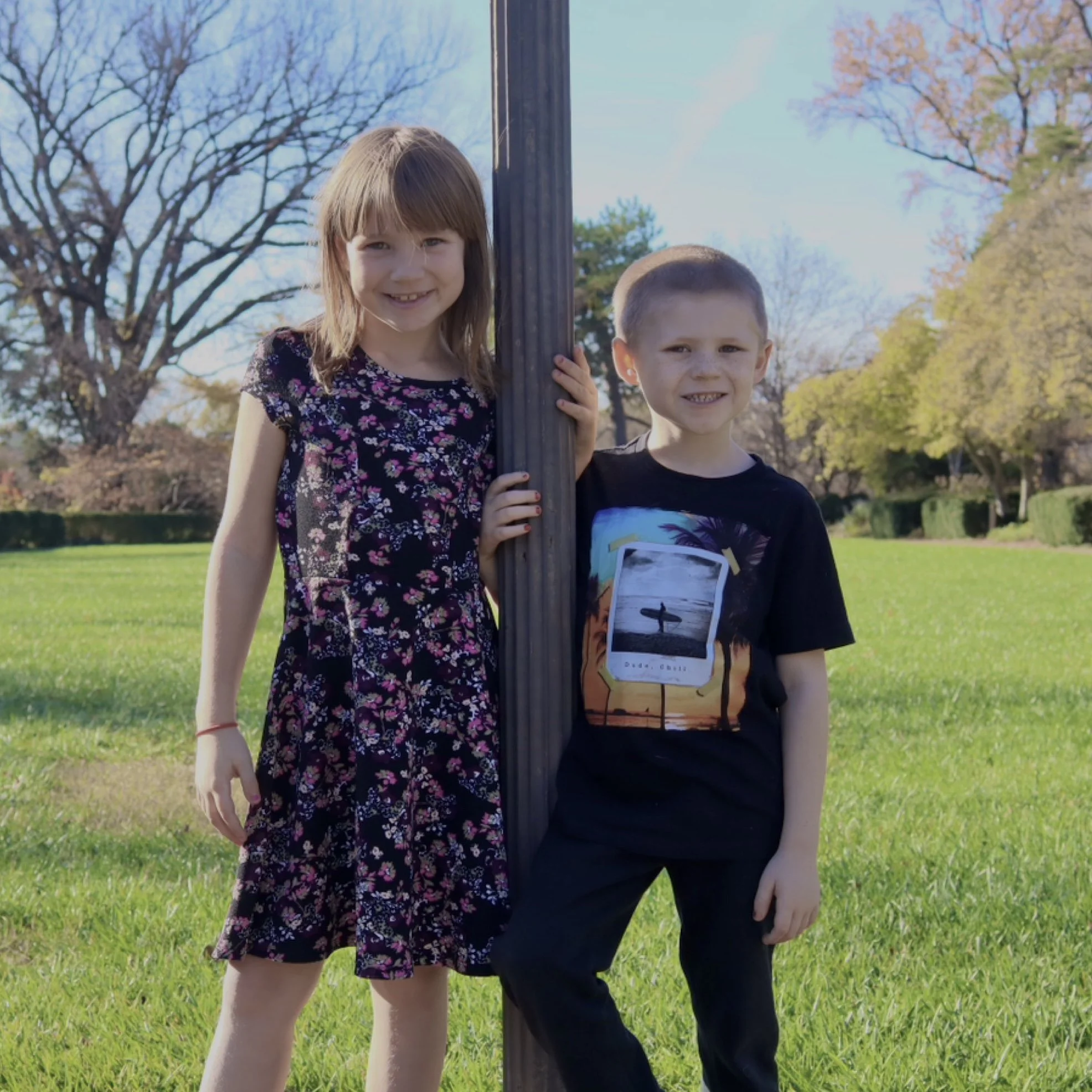 Two children, a girl and a boy, stand by a lamppost in a park, smiling at the camera, with trees and a grassy area in the background during daytime.