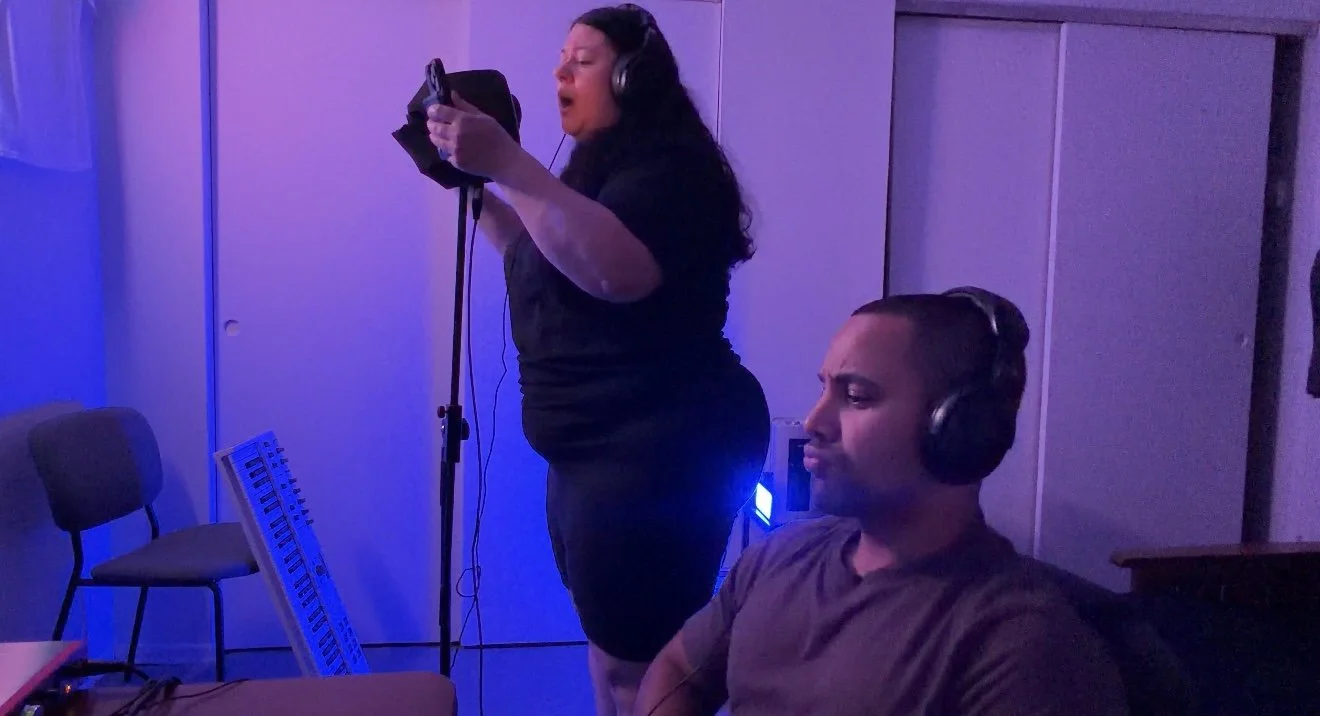A woman singing into a microphone with headphones on, standing, in a recording studio, and a man sitting nearby wearing headphones, in a room illuminated with blue and purple lighting.