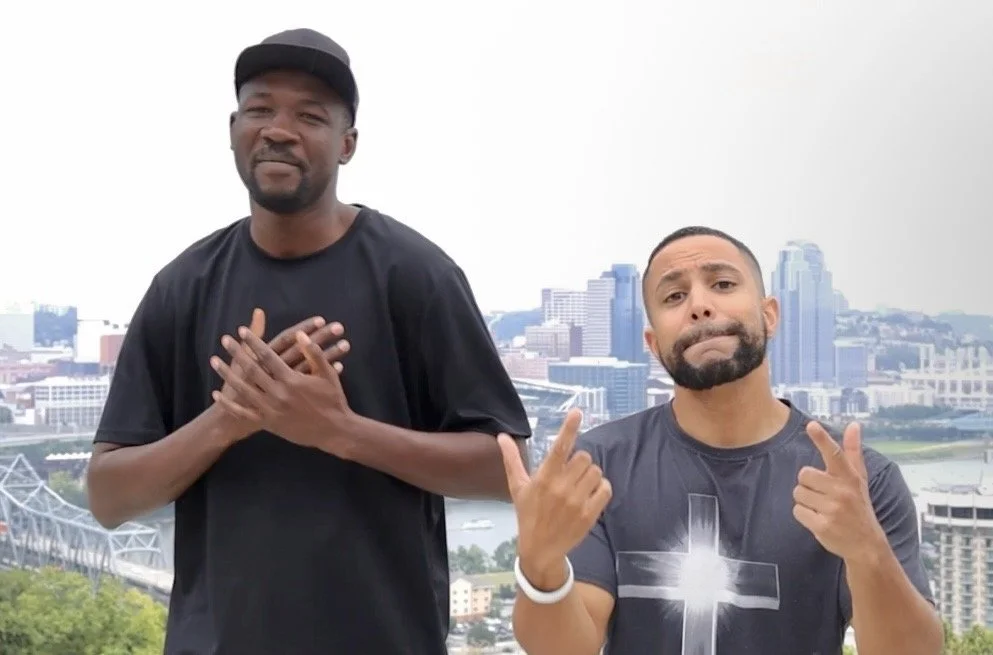Two men standing outdoors with a cityscape background, making expressive gestures and faces.